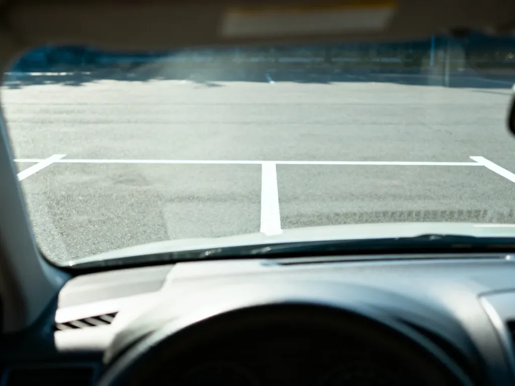 Widok z wnętrza samochodu na puste miejsce parkingowe wyznaczone białymi liniami, przygotowane do manewru parkowania.